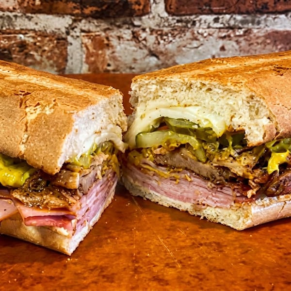Large sandwich, cut in half, revealing layers of ham, roasted meat, melted cheese, pickles, and mustard.