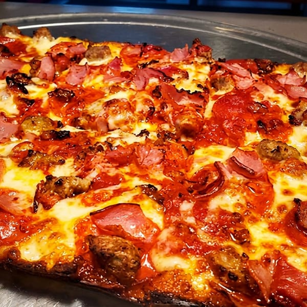 Loaded rectangular pizza with pepperoni, sausage, and ham on a metal tray.